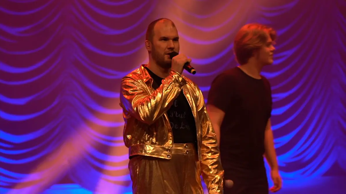 Man i guldfärgad kostym sjunger in i mikrofon på scen. I bakgrunden syns en man i svart tröja.