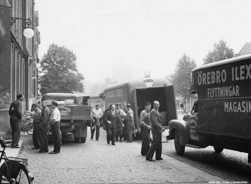 Män i arbetskläder framför tre flyttbilar, parkerade framför ett stort hus.