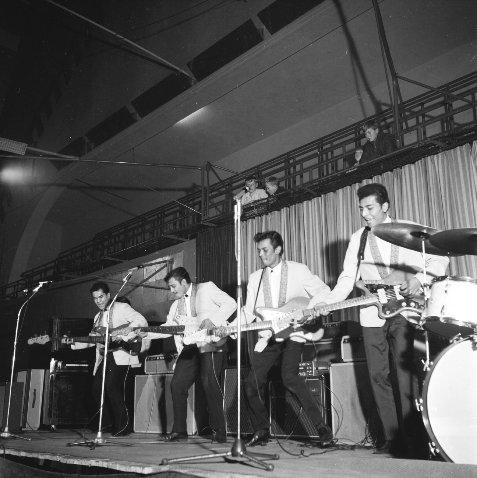 Fyra unga män i likadana ljusa kavajer och frisyrer på en scen. De tre närmast kameran spelar elgitarr, mannen längst bort spelar elbas.