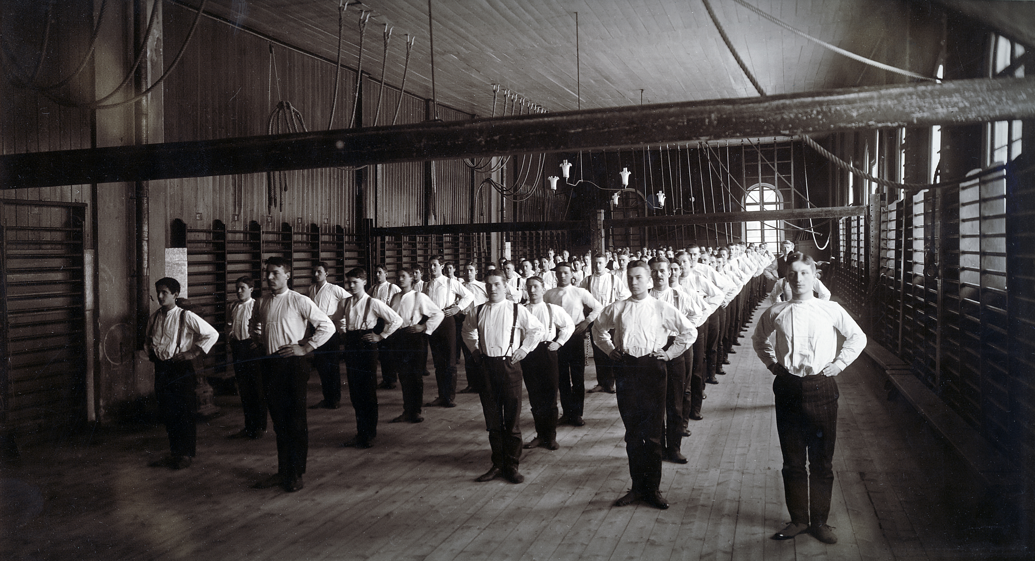 Unga män i vita skjortor och mörka byxor står i fem långa rader. Alla står rakt med armarna på höfterna. Trägolv, bommar, ribbstolar och rep syns i rummet.