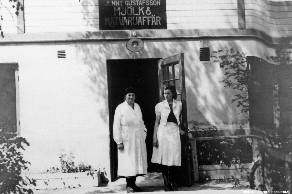 Två kvinnor i ljusa kläder står i en dörr. Över dörren en skylt: "Jenny Gustafsson Mjäölk och matvaruaffär".