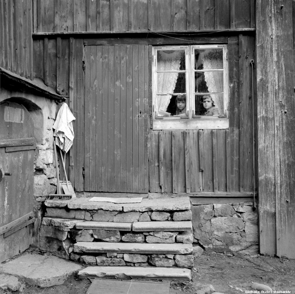 Stentrappa leder upp till trähus. Två barn kikar ut genom ett fönster i huset.