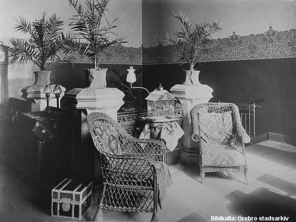 Trappavsats möblerad med två korgstolar, bord med fågelbur och stora växter.