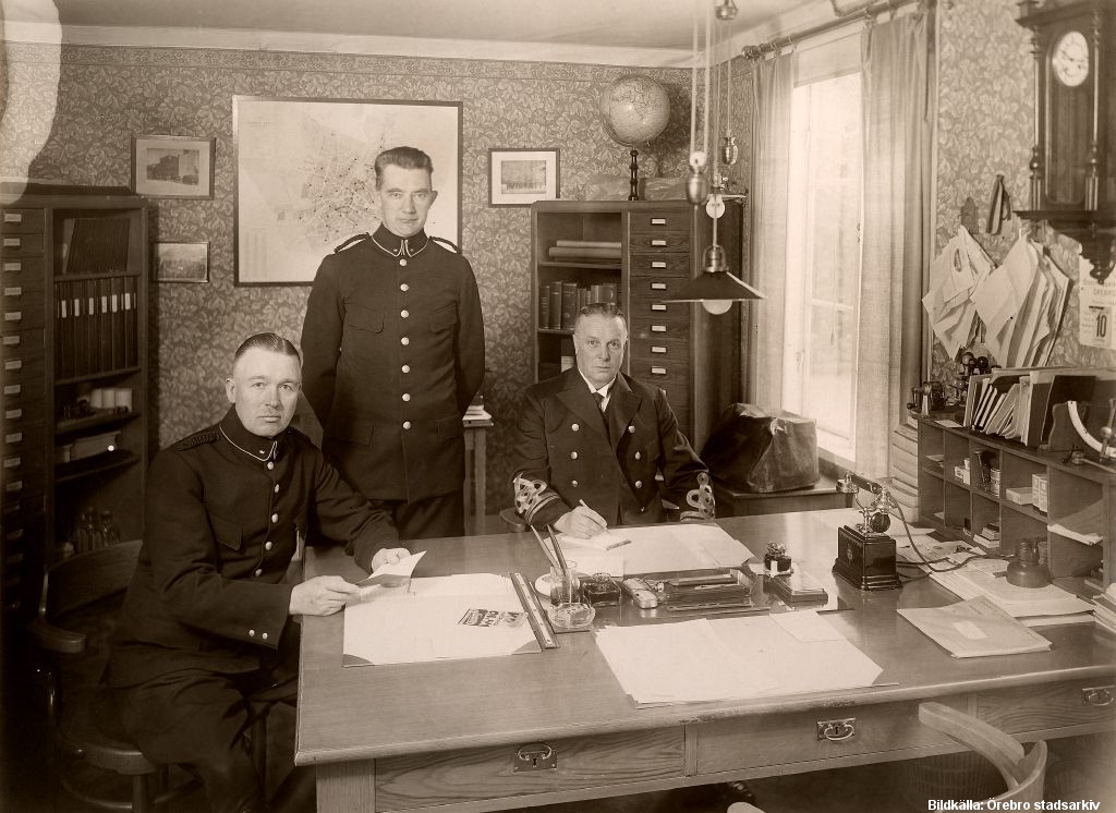 Tre män i uniform bakom ett stort skrivbord inne på ett kontor.
