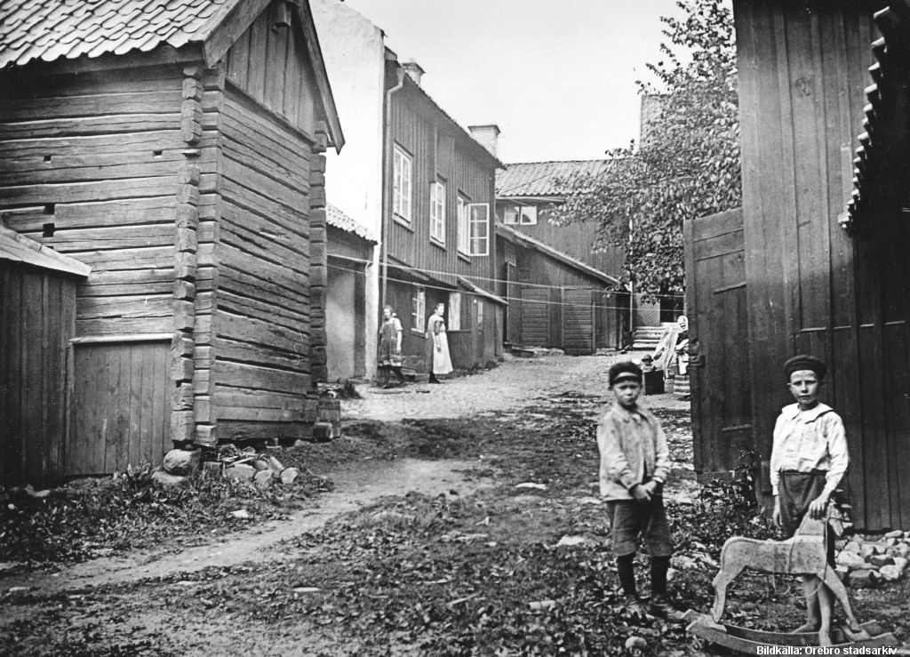 Två barn och en liten gunghäst står på en bakgård med trähus. 