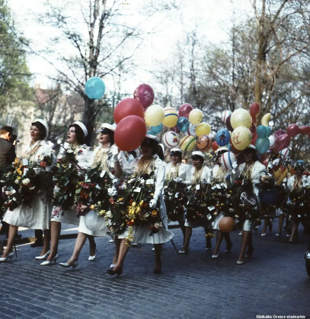 Unga kvinnor i vita klänningar och vita mössar går i formation. Alla har blommor runt halsen, de flesta håller ballonger.